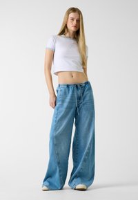 Weißes, kurz geschnittenes T-Shirt mit hellblauen Abschlüssen, kombiniert mit locker sitzenden hellblauen Jeanshosen mit Kordelzug in der Taille und weiten Beinen.