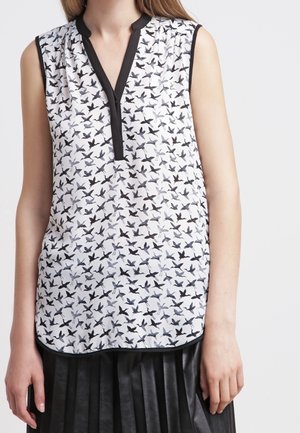 Ærmeløs hvid bluse med sort fugleprint og sort kant på krave og slids, båret med sort plisseret nederdel.