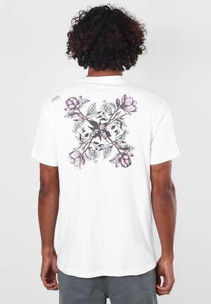 Homme aux cheveux bouclés portant un t-shirt blanc avec un grand motif floral violet et feuilles dans le dos.