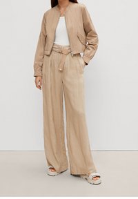 Beige cropped bomberjakke med lynlås, parret med højtaljede bredbenede bukser med en stofbælte. Begge dele er lavet af letvægtsmateriale.