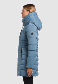Giacca puffer blu con colletto alto e cappuccio. Presenta una texture trapuntata, tasche laterali e una toppa con logo sulla manica. Design a taglio dritto.