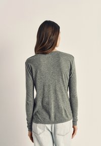 Moterys su ilgais rudos spalvos plaukais, žvelgiančios į šoną, dėvinčios pilką ilgomis rankovėmis marškinius ir šviesiai mėlynas džinsines kelnes prieš paprastą šviesią foną.