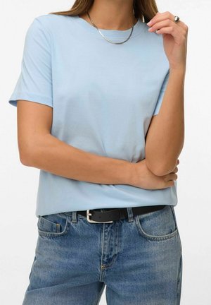 Femme portant un t-shirt bleu clair à manches courtes rentré dans un jean bleu avec une ceinture noire, un collier en argent et une bague à la main droite.