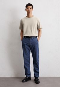 Beige Baumwoll-T-Shirt mit kurzen Ärmeln, kombiniert mit dunkelblauen strukturierten Hosen und schwarzen Lederschuhen, versehen mit einem einfachen Gürtel.
