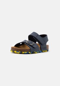 Sandalias azules con dos correas ajustables, una hebilla y una plantilla de corcho. La suela presenta un diseño colorido y con patrón en amarillo y azul.