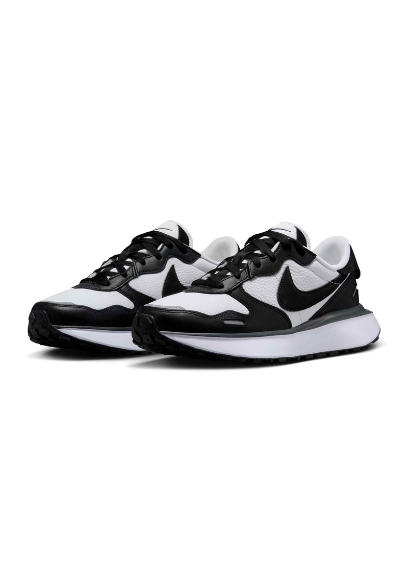 Zapatillas deportivas en blanco y negro con parte superior de malla y cuero, presentan una suela acolchada, diseño con cordones y un logo prominente en el lateral.