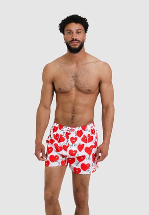 Shirtless man with curly hair and beard wearing white boxer shorts patterned with red cartoon hearts on a plain background.