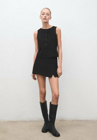 Top senza maniche nero con bottoni sul davanti, abbinato a una mini gonna nera con taglio diagonale e stivali neri al ginocchio. Design minimalista.