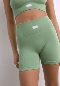 Prsni športni komplet svetlo zelene barve z rebrasto teksturo, ki vključuje kratko majico in kratke hlače z visokim pasom. Material je raztegljiv; na vsakem kosu sta majhni oznaki z logotipom.