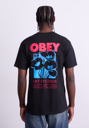 Black cotton t-shirt featuring a colorful graphic on the back with the words "OBEY" in red and "ART STUDIOS" in red below, and floral design.