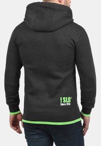 Mørkegrå hoodie med hætte, grøn kantbånd og hvidt logo nederst på ryggen. Fremstillet af et blødt, struktureret stof.