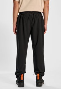 Czarne spodnie sportowe z elastycznym paskiem w talii, dopasowanym krojem i sznurkiem przy kostkach. Noszone z czarnymi butami z pomarańczowymi akcentami.