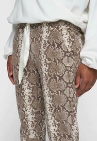 Bruine en crèmekleurige slangenprint broek met een gladde textuur, elastische tailleband en witte satijnen koord met metalen uiteinden.