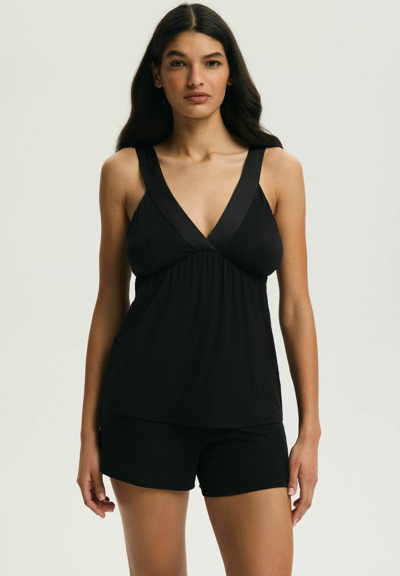 Žena s dlhými tmavými vlasmi, oblečená v čiernom bezrukávovom topu s výstrihom do V a čiernych krátkych nohaviciach, stojaca pred jednoduchým svetlým pozadím.