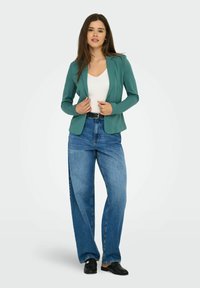 Blazer en teal à coupe slim, tissu texturé, haut blanc ajusté, jeans taille haute bleu clair et chaussures noires à enfiler.