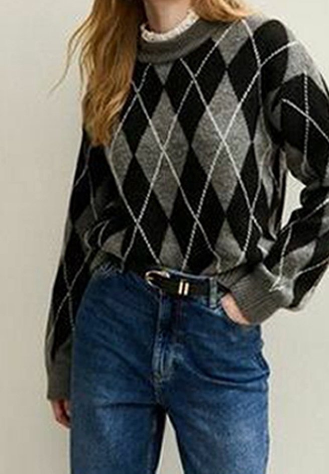 Femme portant un pull gris et noir à motif losange, un jean bleu, une main dans la poche, et une ceinture noire avec une boucle dorée.
