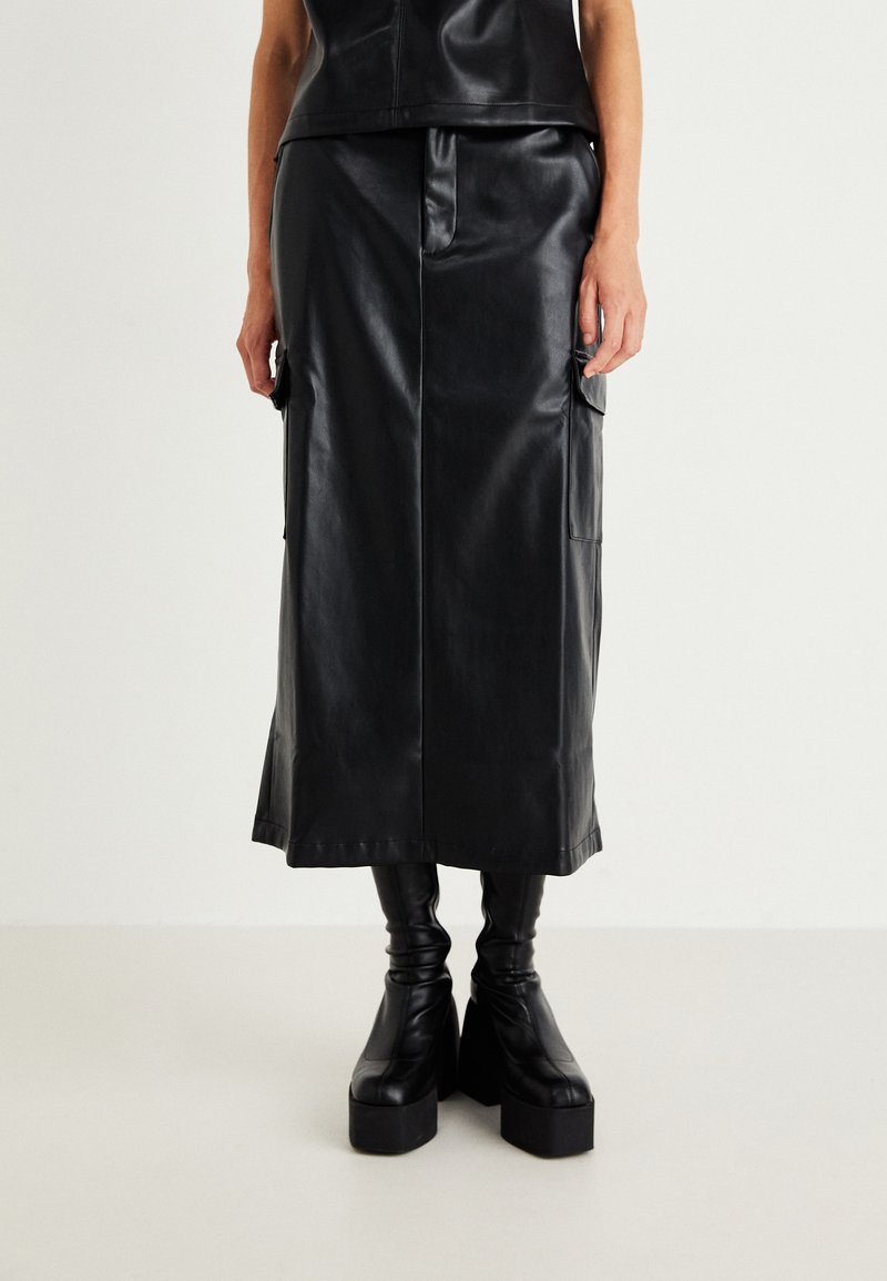 Schwarzer Leder-Midirock mit geradem Design, ausgestattet mit zwei seitlichen Taschen und einer glatten Textur. Kombiniert mit klobigen schwarzen Plateau-Stiefeln.