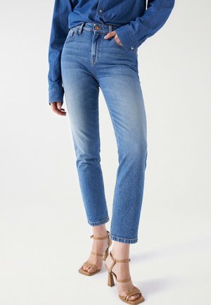 Vrouw met lichtblauwe rechte jeans met hoge taille, een spijkerblouse en beige hoge hakken sandalen, staand tegen een witte achtergrond.