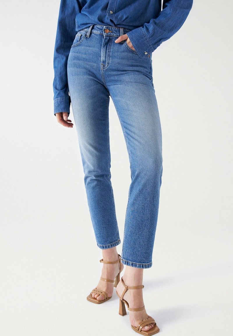 Femme portant un jean droit taille haute bleu clair, une chemise en denim et des sandales à talons hauts couleur beige, debout sur fond blanc.