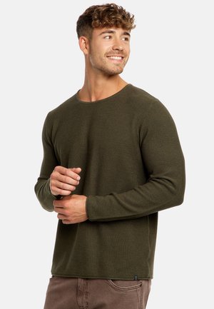 Joven con pelo rizado que lleva un jersey de manga larga verde oliva y pantalones marrones, mirando hacia un lado y sonriendo.