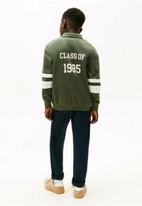 Groene sweatshirt met witte strepen, "CLASS OF 1985" gedrukt op de achterkant. Gecombineerd met marineblauwe broek en lichtgekleurde schoenen.