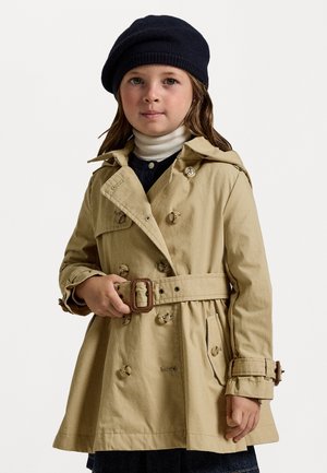 Beige trenchcoat med dubbelknäppta knappar, ett bälte i midjan och axelklaffar, i kombination med en marinblå basker och en vit polotröja.