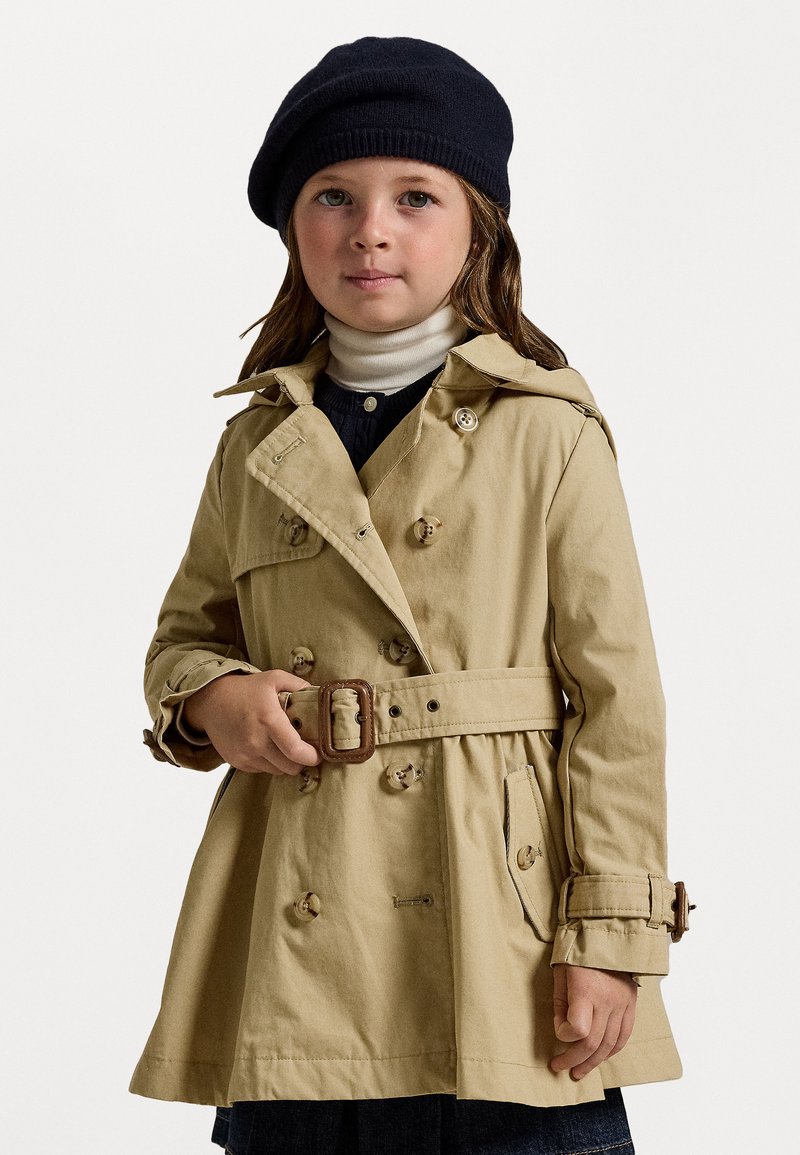 Beige trenchcoat med dubbelknäppta knappar, ett bälte i midjan och axelklaffar, i kombination med en marinblå basker och en vit polotröja.
