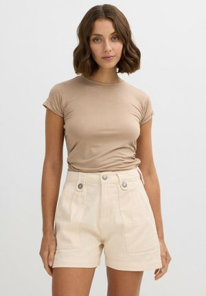 Camiseta ajustada de color beige claro con mangas enrolladas, combinada con pantalones cortos de crema claros de cintura alta que cuentan con dos bolsillos frontales y dos botones metálicos.