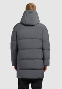 Muškarac u dugom sivoj podstavljenoj kaputu s velikim kapuljačom, prikazan sa leđa na jednostavnoj pozadini.