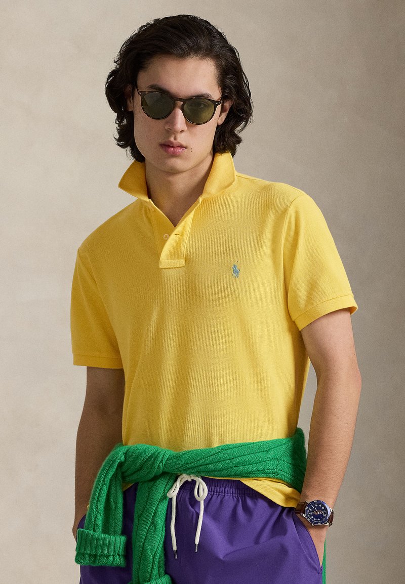 Κίτρινη κοντομάνικη πόλο μπλούζα με ριμπ γιακά, διακοσμημένη με μικρό μπλε λογότυπο. Φορεμένη με πράσινο πουλόβερ δεμένο στη μέση και μωβ σορτς.