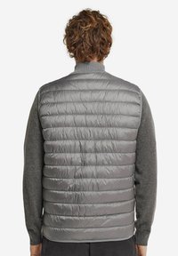 Graue Steppweste mit gestepptem Design und hohem Kragen, über einem dunkelgrauen Langarmshirt getragen. Glatte, glänzende Textur.