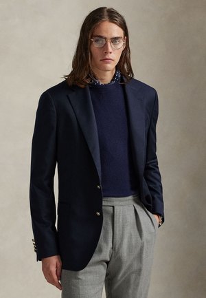 Jeune personne portant des lunettes rondes, un blazer bleu marine, un pull bleu marine, un col de chemise à pois et un pantalon gris plissé taille haute, les mains dans les poches.