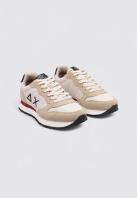 Baskets beige et crème avec tige en daim et en mesh, bout rond, devant à lacets et accent rouge sur la semelle. Logo noir sur la languette.