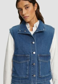 Vrouw in een blauwe denim knoopvest met klepzakken over een witte blouse met lange mouwen, tegen een lichte achtergrond.