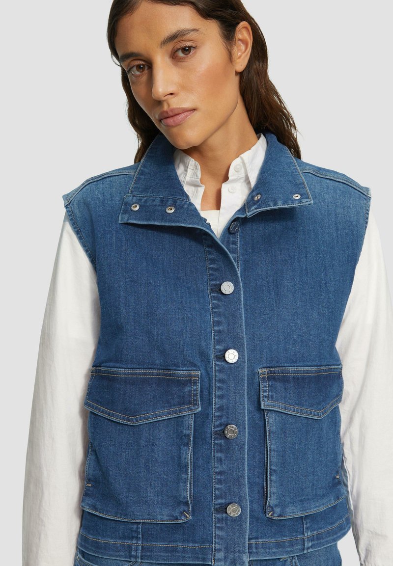 Vrouw in een blauwe denim knoopvest met klepzakken over een witte blouse met lange mouwen, tegen een lichte achtergrond.
