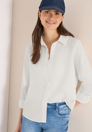 Femme souriante portant une chemise blanche texturée à boutons, un jean en denim bleu et une casquette de baseball bleu foncé, sur un fond neutre.