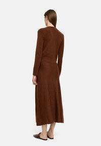 Brązowa dzianinowa sukienka dwuczęściowa z długimi rękawami, dopasowanym gorsetem i zwiewną spódnicą. Teksturowany materiał; noszona z brązowymi klapkami.