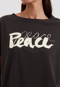 Schwarzes T-Shirt mit dem Wort "Frieden" in weißer, ausdrucksvoller Schrift. Kurze Ärmel und ein weiter Ausschnitt. Aus weichem Baumwollstoff.