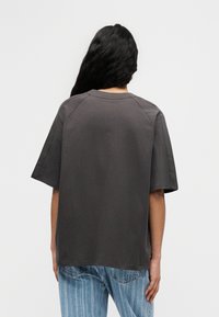 Άτομο με μακριά σκούρα μαλλιά, που φοράει άνετο σκούρο γκρι μπλουζάκι και ανοιχτό μπλε τζιν με ρίγες, γυρισμένο πλάτη σε απλό λευκό φόντο.