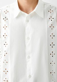 Bílá košile na knoflíky s propracovanou výšivkou z dírkované krajky podél stran, jemnou texturou a klasickým límečkem.