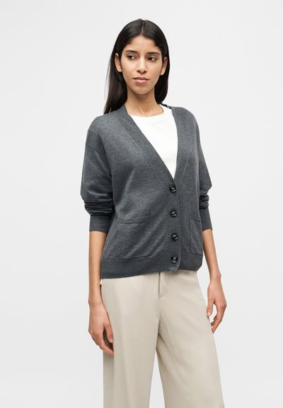 Γκρι πλεκτή ζακέτα με V-λαιμόκοψη, μακριά μανίκια, πέντε κουμπιά και δύο μπροστινές τσέπες, φορεμένη πάνω από λευκή μπλούζα, σε συνδυασμό με μπεζ παντελόνια.