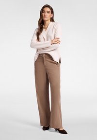 Femme debout les bras croisés, portant un pull côtelé rose clair, un pantalon large marron, un collier en or et des escarpins pointus foncés.
