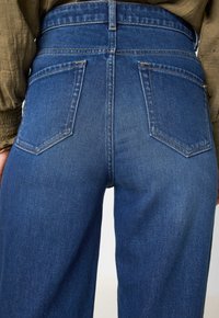 Jeans en denim bleu taille haute avec un design classique à cinq poches, une texture légère et des coutures visibles sur les poches et la ceinture.