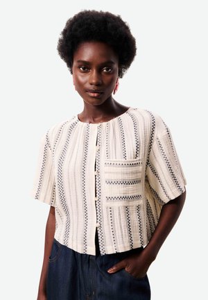 Femme portant un chemisier crème à manches courtes avec un motif à rayures verticales noires et des boutons, associé à un pantalon bleu foncé, main dans la poche.