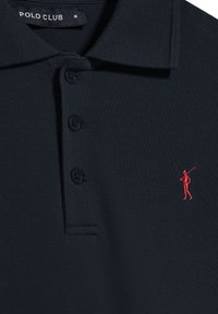 Marinblå polo med texturerat tyg, treknapplås och en röd broderad logotyp av en golfspelare på vänster bröst. Stl M.