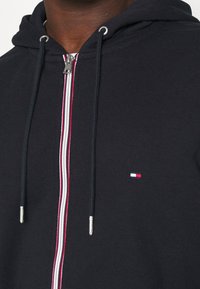 Felpa nera con cappuccio e zip argentata, con dettagli a strisce rosse e bianche sul davanti e un piccolo logo sul petto sinistro.