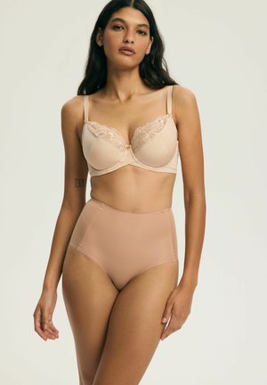Kobieta z długimi ciemnymi włosami, ubrana w beżowy biustonosz z koronką i beżową modelującą bieliznę wysokiego stanowi na jasnym tle.