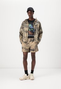 Leichte, beigefarbene Jacke mit einem Naturdruck, schwarzes T-Shirt mit Grafik, passende Shorts und cremefarbene Sneakers; kombiniert mit einer schwarzen Mütze und schwarzen Socken.