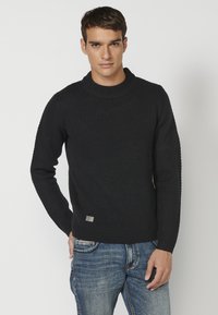 Schwarzer Strickpullover mit strukturiertem Muster und rundem Ausschnitt. Verfügt über ein kleines Logopatch an der unteren linken Seite. Getragen über blauen Jeans.