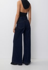 Femme portant un jean large noir et un haut sans manches et dos nu noir, se tenant devant un fond clair uni.